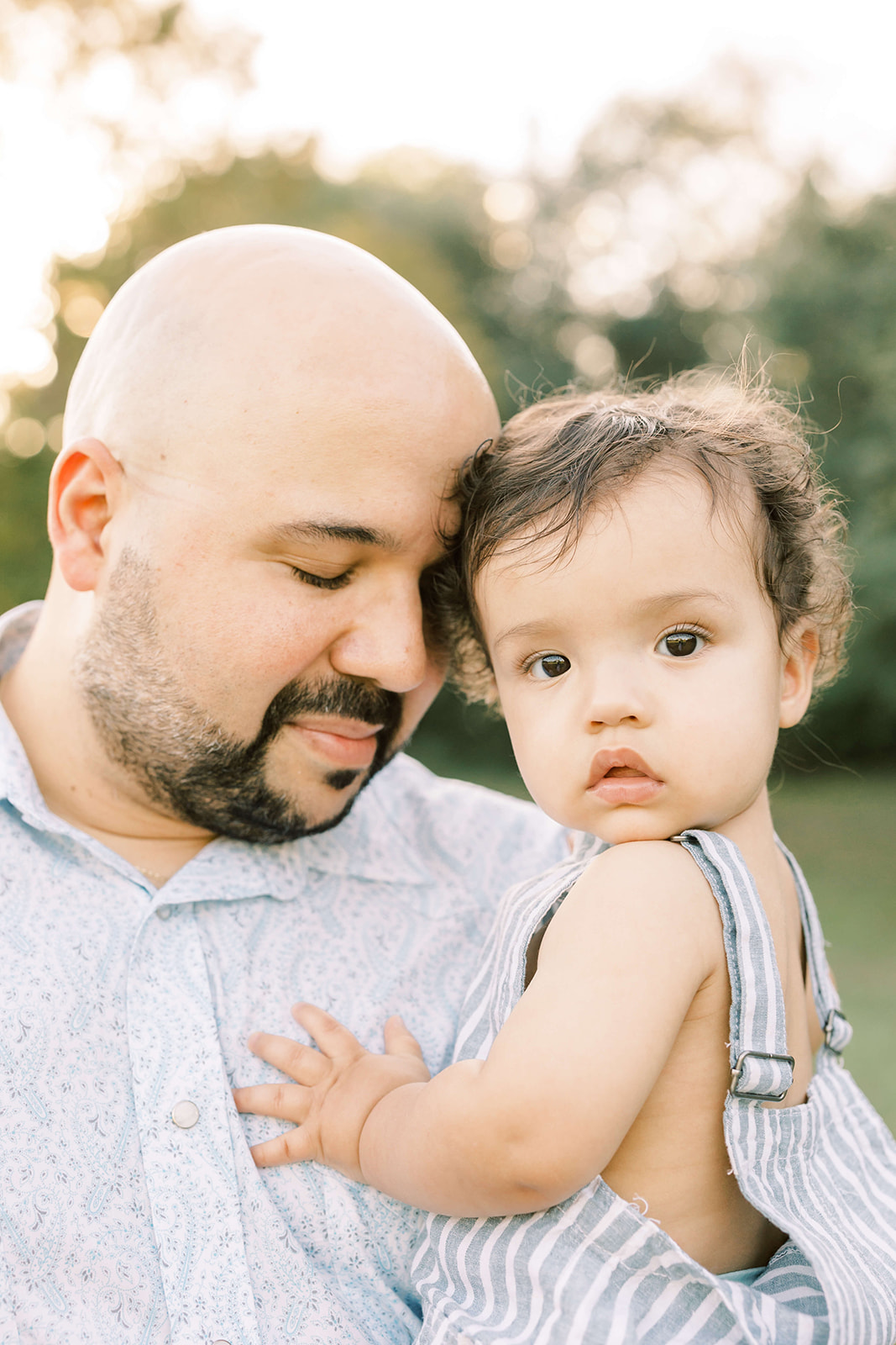 A father touches foreheads with his toddler son in overalls on his hip before visiting Houston pediatric dentists