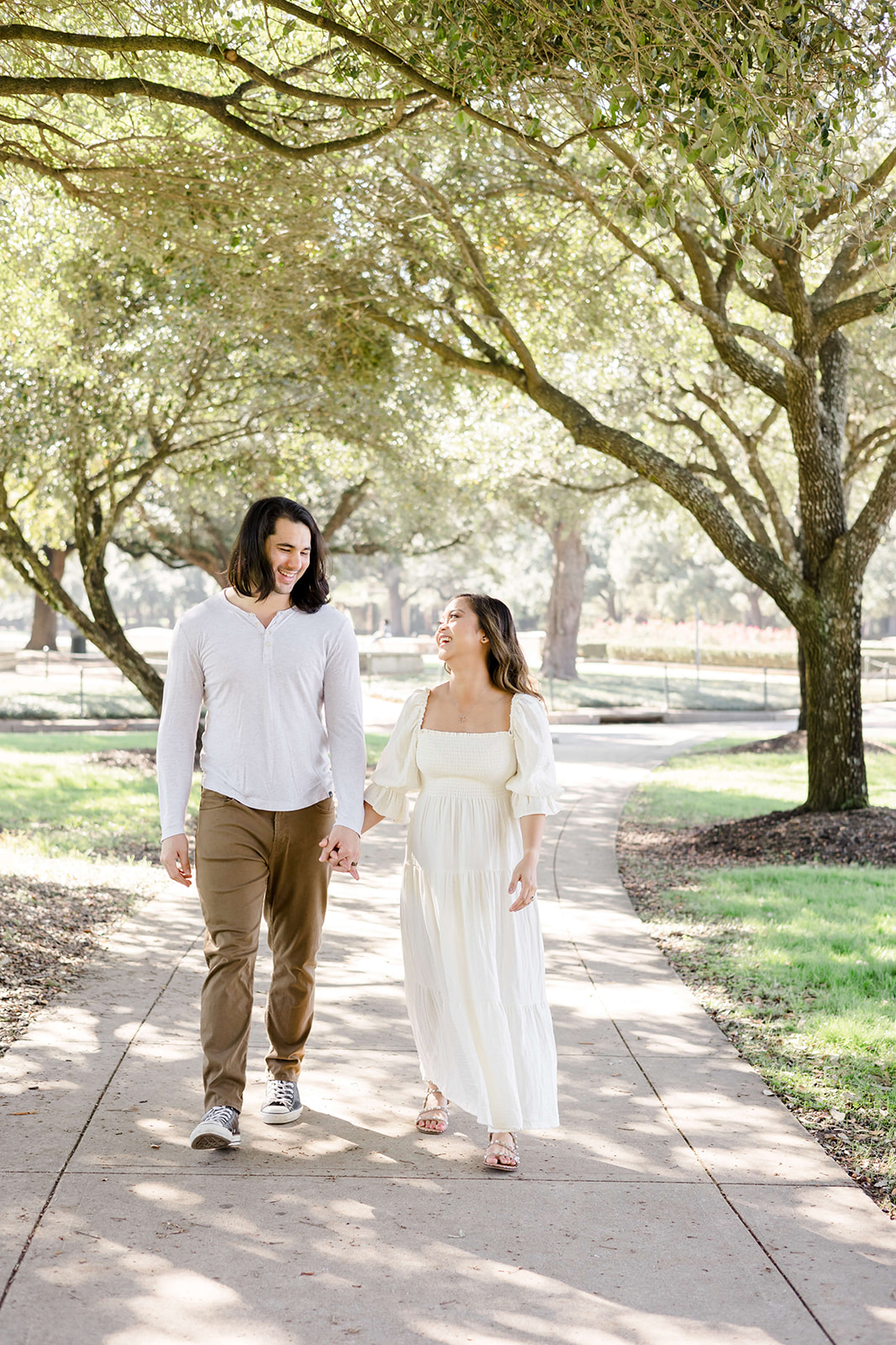 Happy expecting parents walk hand in hand down a park sidewalk laughing at sunset