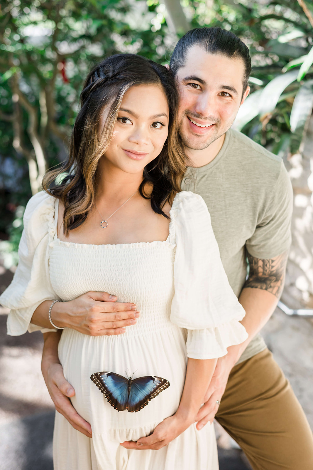 A butterfly sits on the pregnant bump of a mom to be while she stands in a garden with her husband