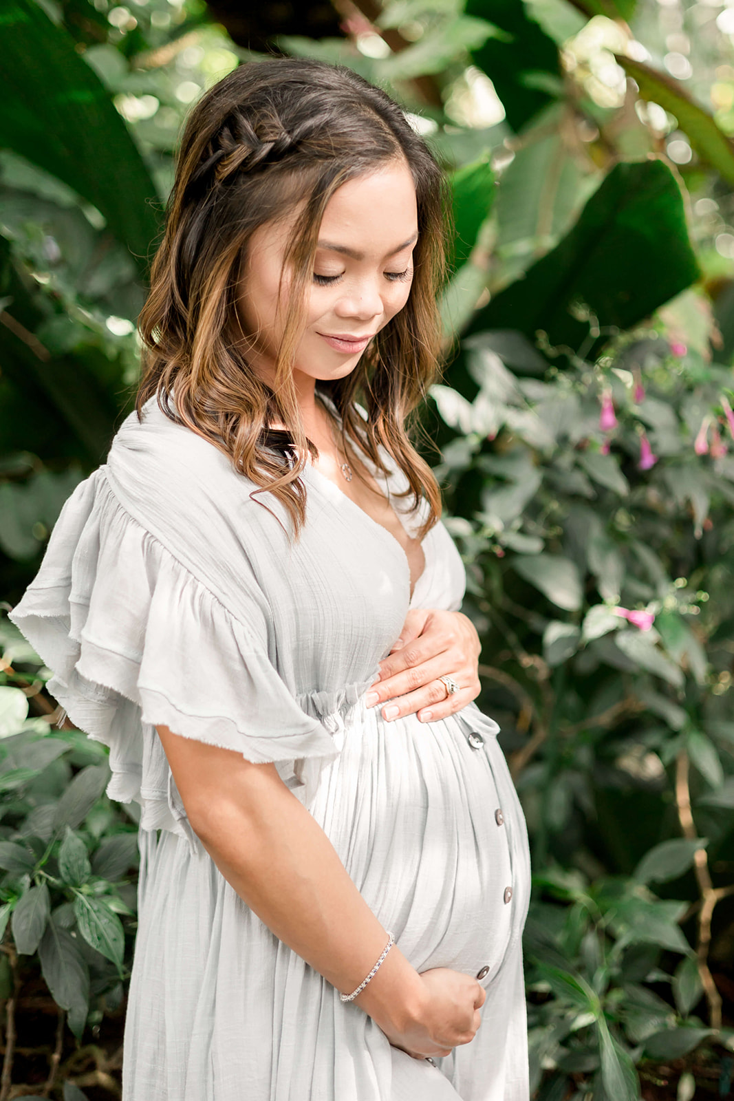 A mom to be in a blue maternity dress smiles down to her bump while standing in a tropical garden before a Houston Prenatal Massage