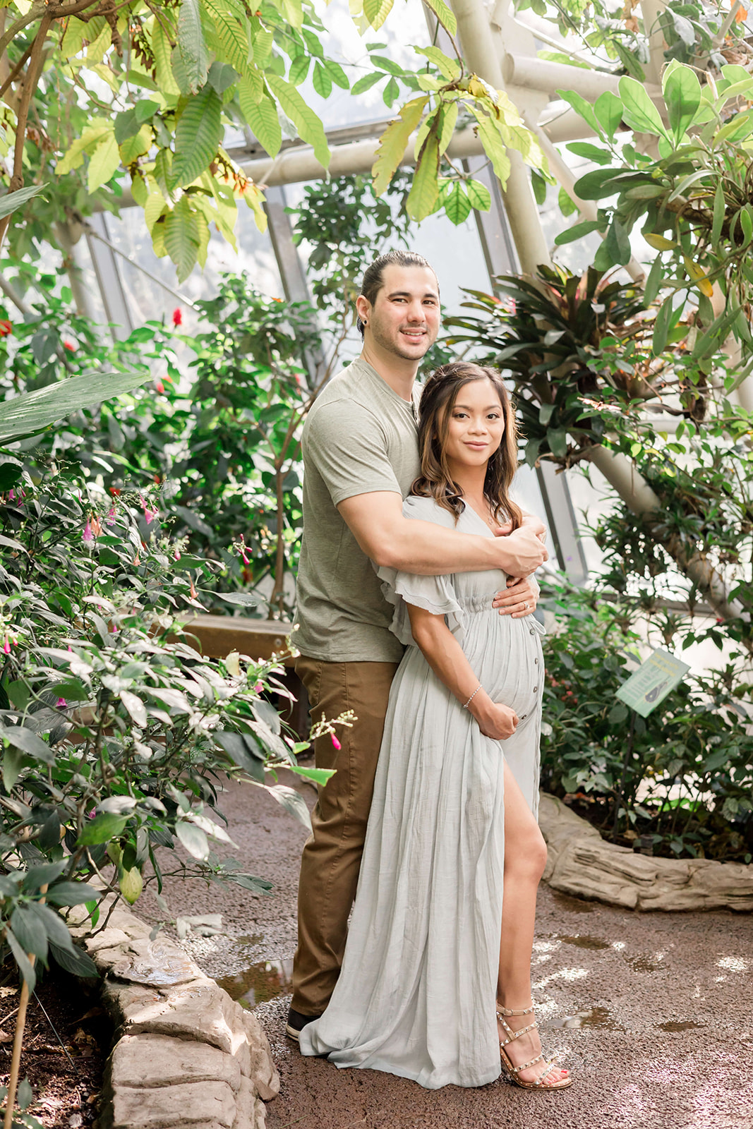 A mom to be stands in the chest of her husband while being hugged in a botanical garden greenhouse before a Houston Prenatal Massage