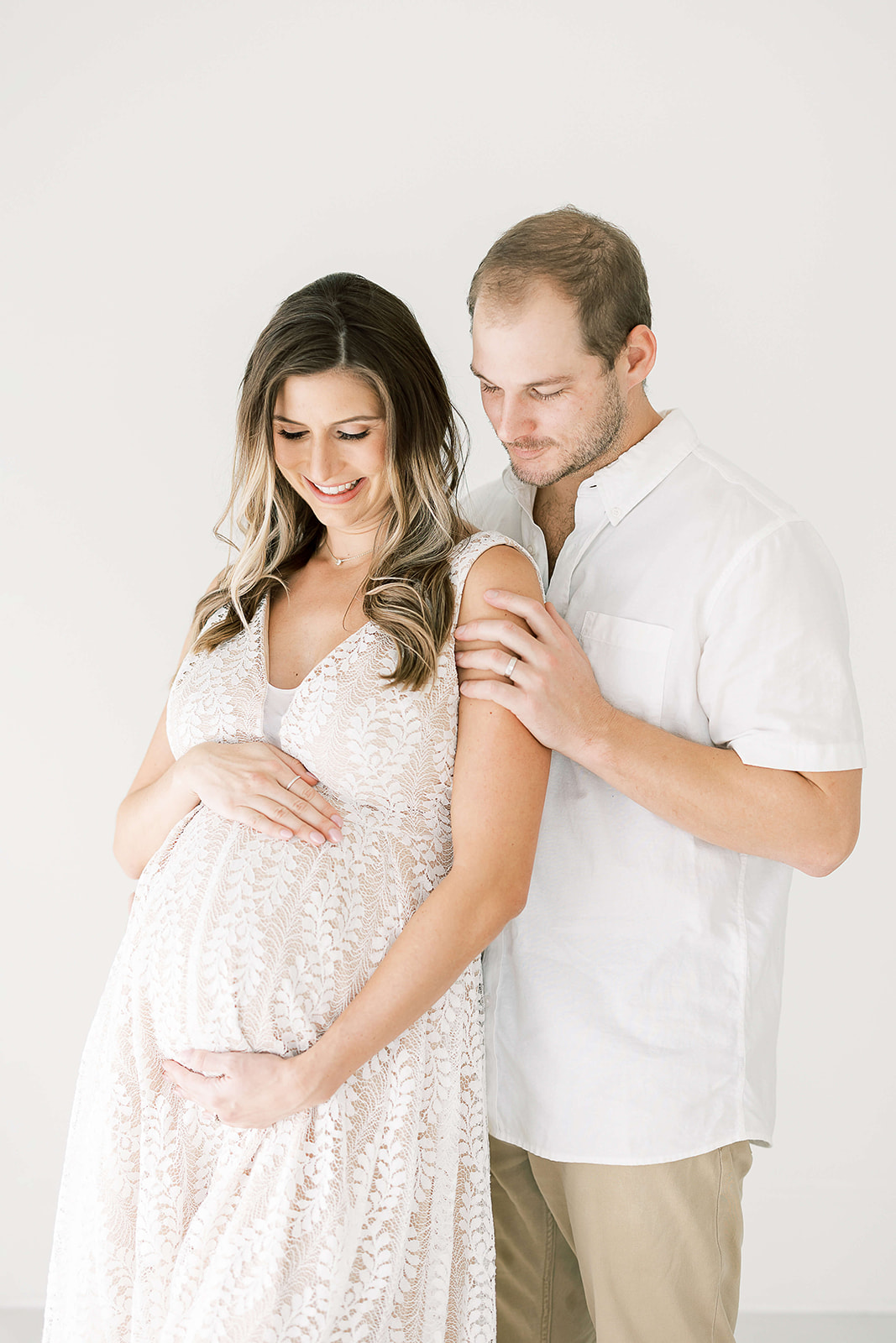 A smiling mom to be leans into the chest of her husband in a studio while they smile at the bump