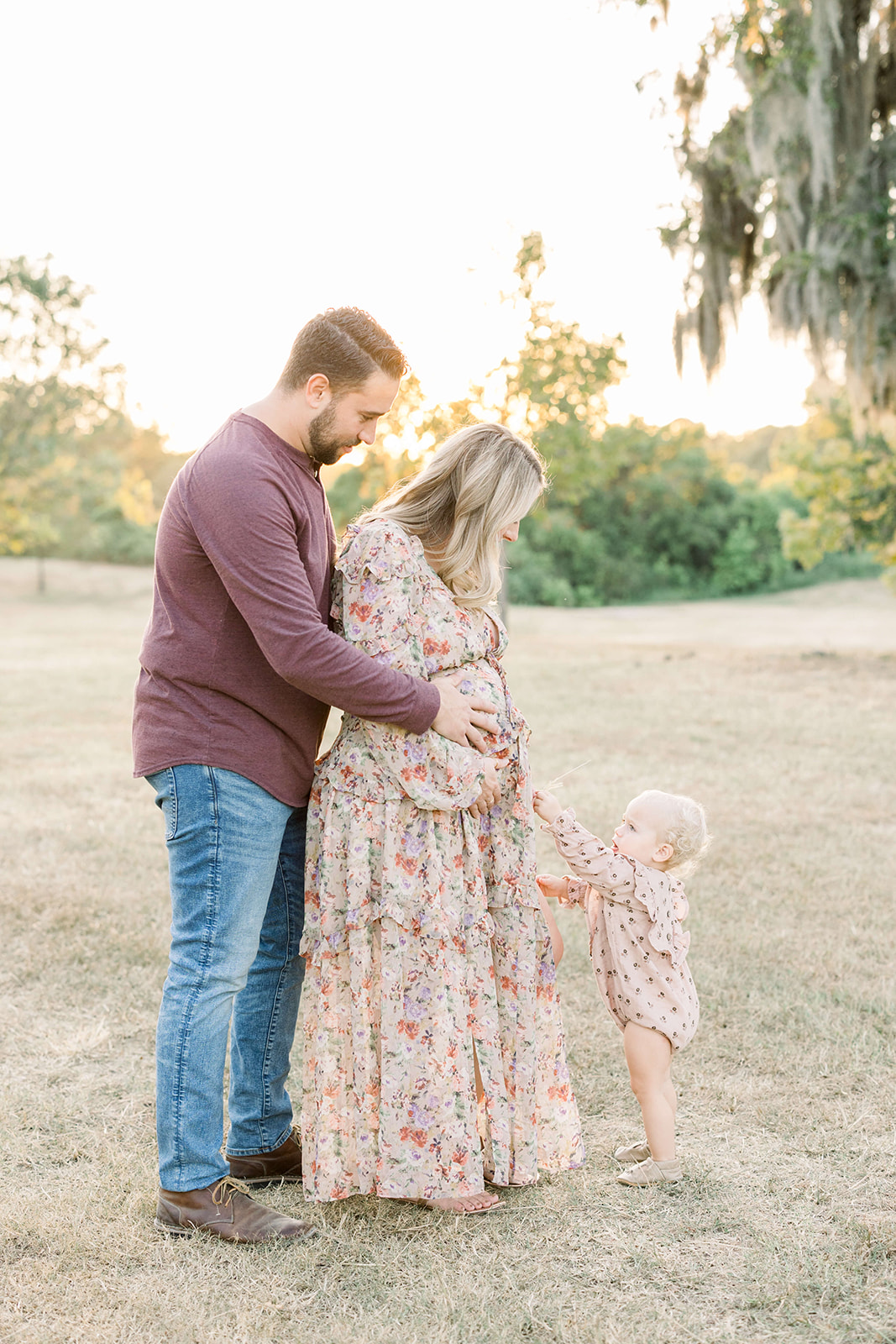 Happy expecting parents stand in a park as their toddler daughter reaches up to the bump