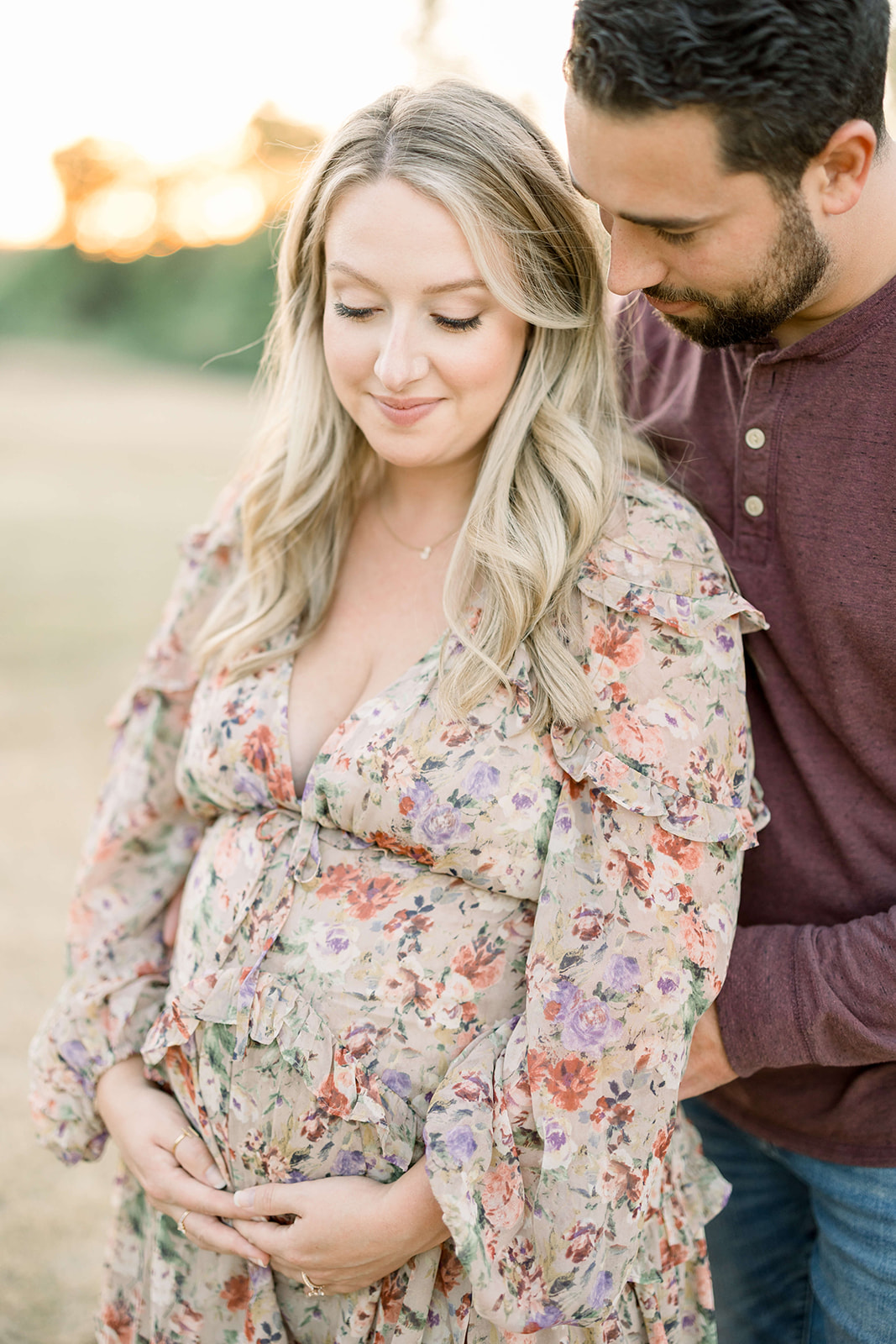 A mom to be in a floral print dress stands in a field at sunset with her husband after visiting Marley And Moo Midwifery