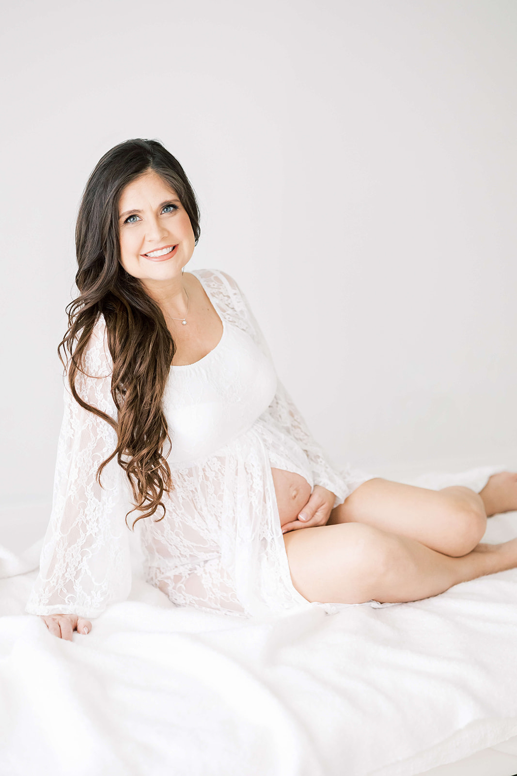 A happy pregnant woman sits across a white bed in a studio while holding the bump