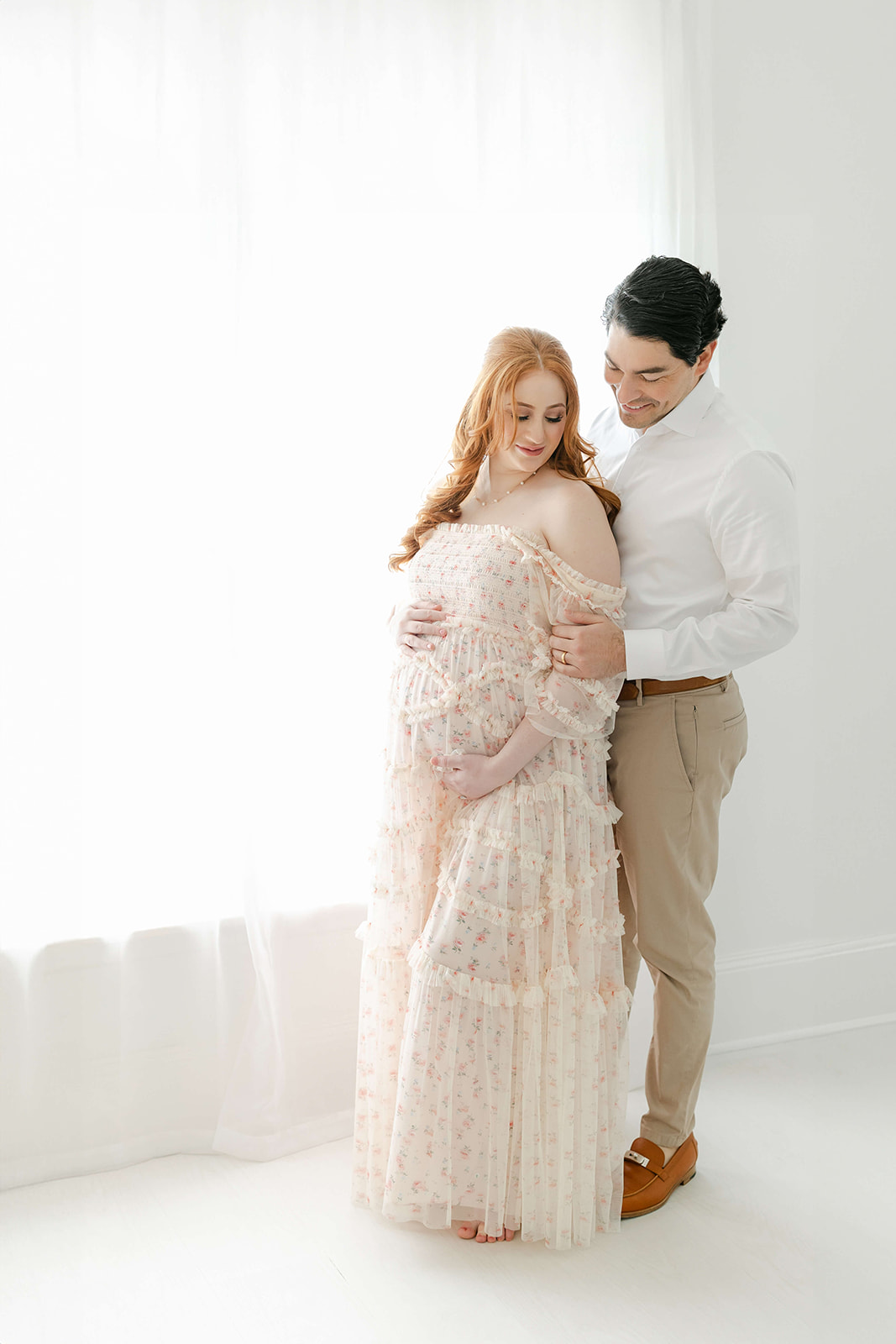 A mom to be leans into the chest of her husband as they stand in a studio holding the bump