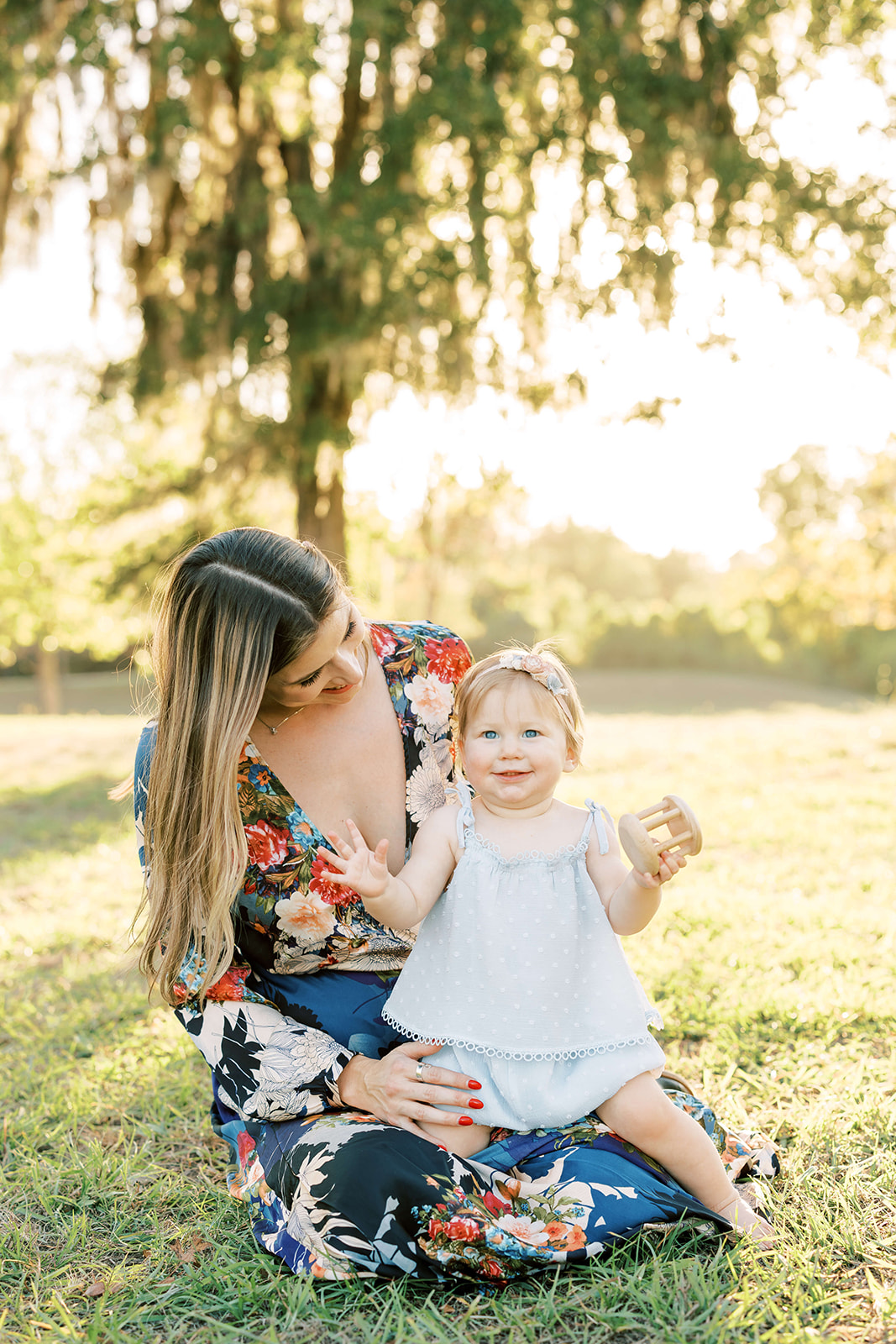 A happy toddler girl plays with a wooden toy in mom's lap in a park at sunset before visiting a Childrens Boutique Houston