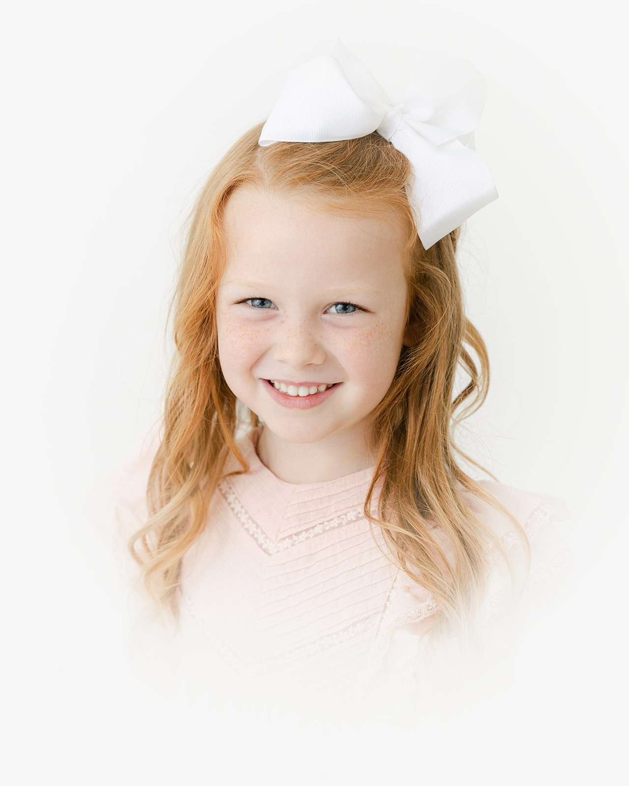 A young girl with red hair wears a large white bow and pink dress in a studio