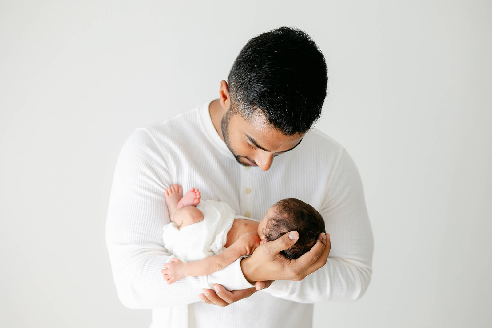 A new father in a white henley smiles down at his newborn baby in his hands before meeting a Houston Au Pair
