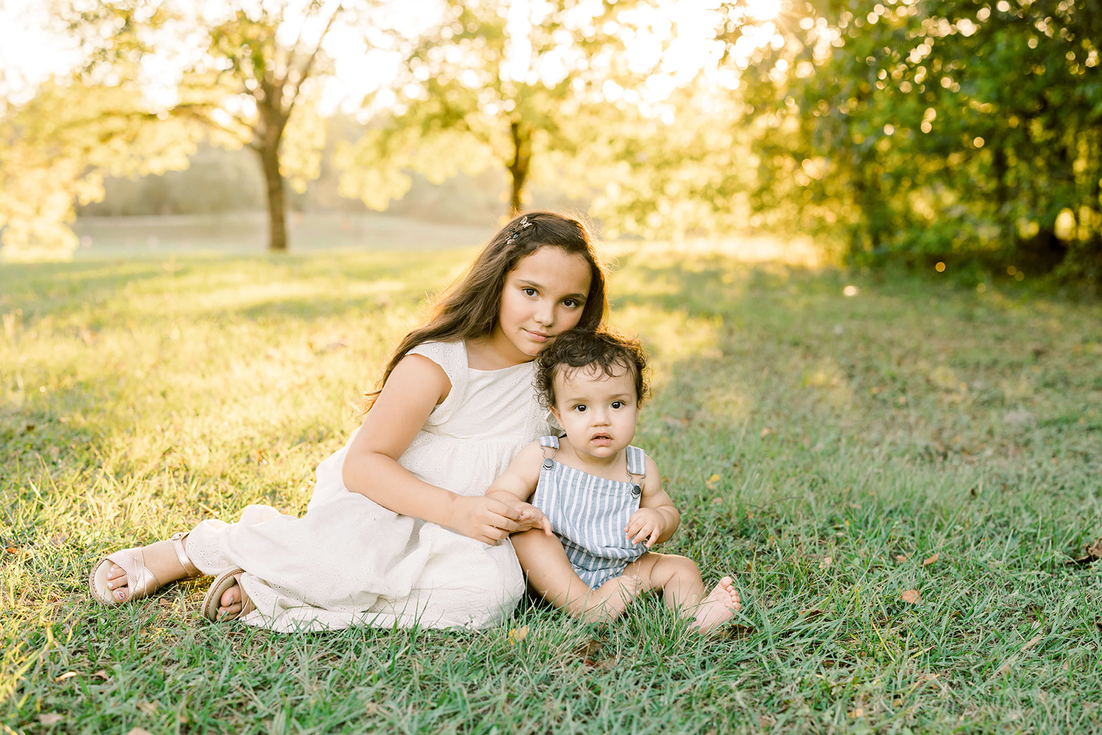 A young girl in a white dress sits in a lawn at sunset with her baby sibling after meeting Houston Babysitters