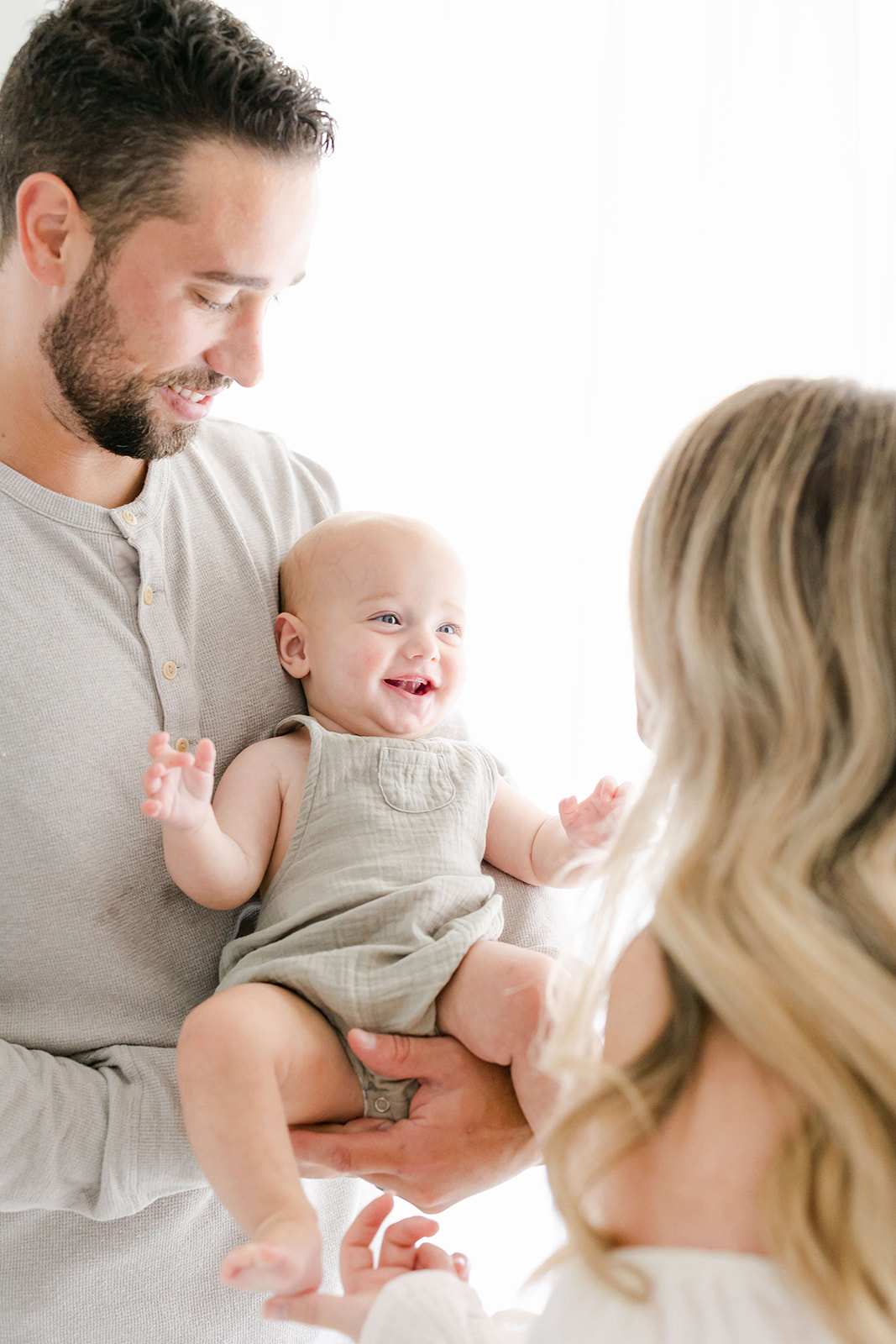 A happy infant boy in green overalls smiles at mom while sitting in dad's arms