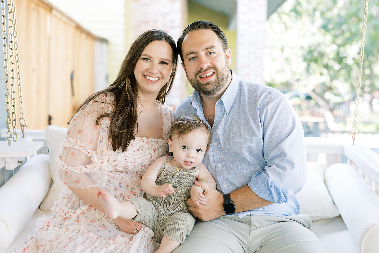 Happy parents sit on a porch swing with their smiling toddler in their lap in a green knit onesie