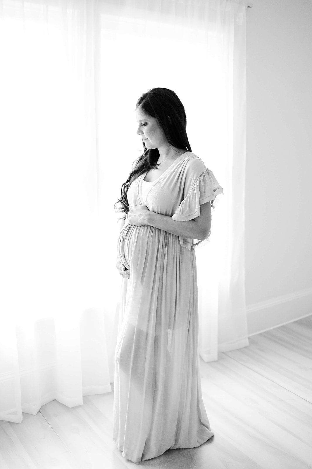 A smiling pregnant woman walks in a studio wearing a white maternity dress and holding her bump