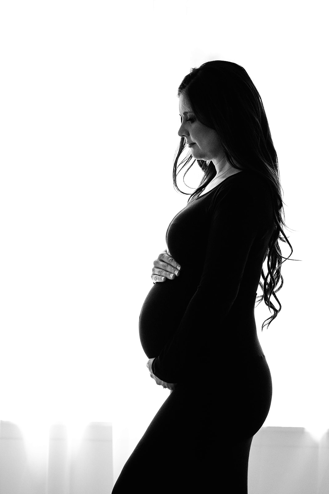 Profile of a mother to be standing in a window holding her bump in a black maternity gown before getting Placenta Encapsulation Houston