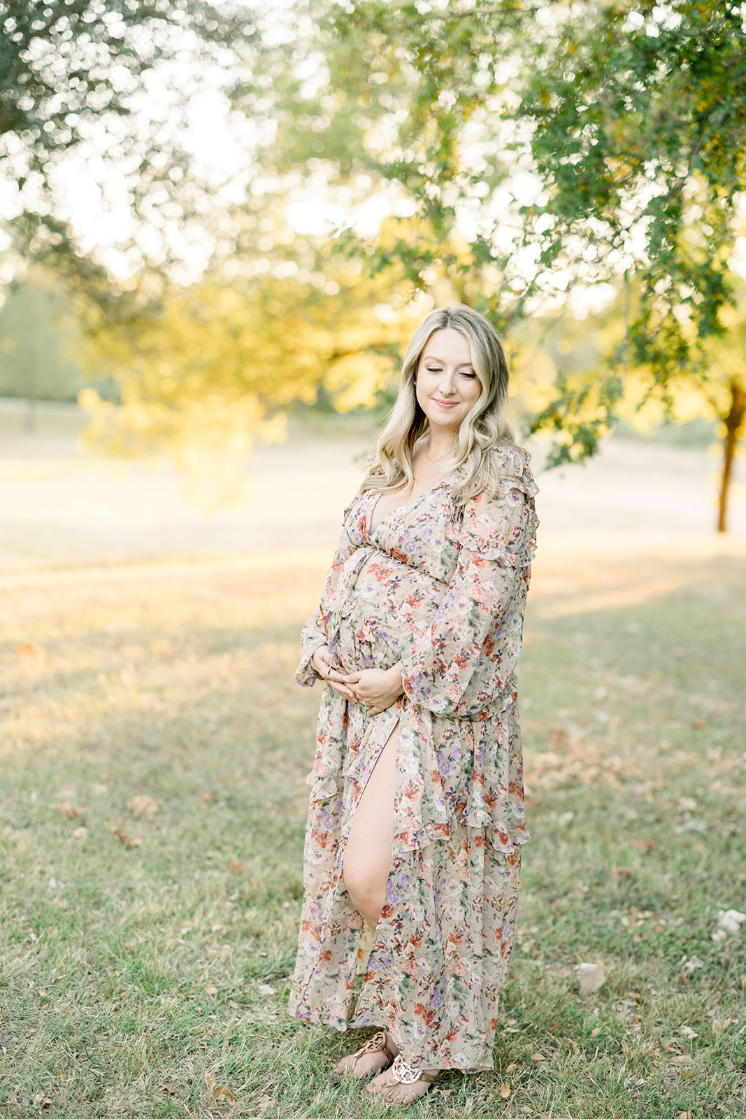 A mother to be in a floral print dress stands in a park lawn at sunset smiling down her shoulder