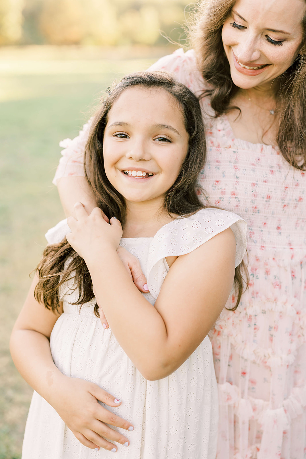 A young girl in a white dress is hugged by mom in a park