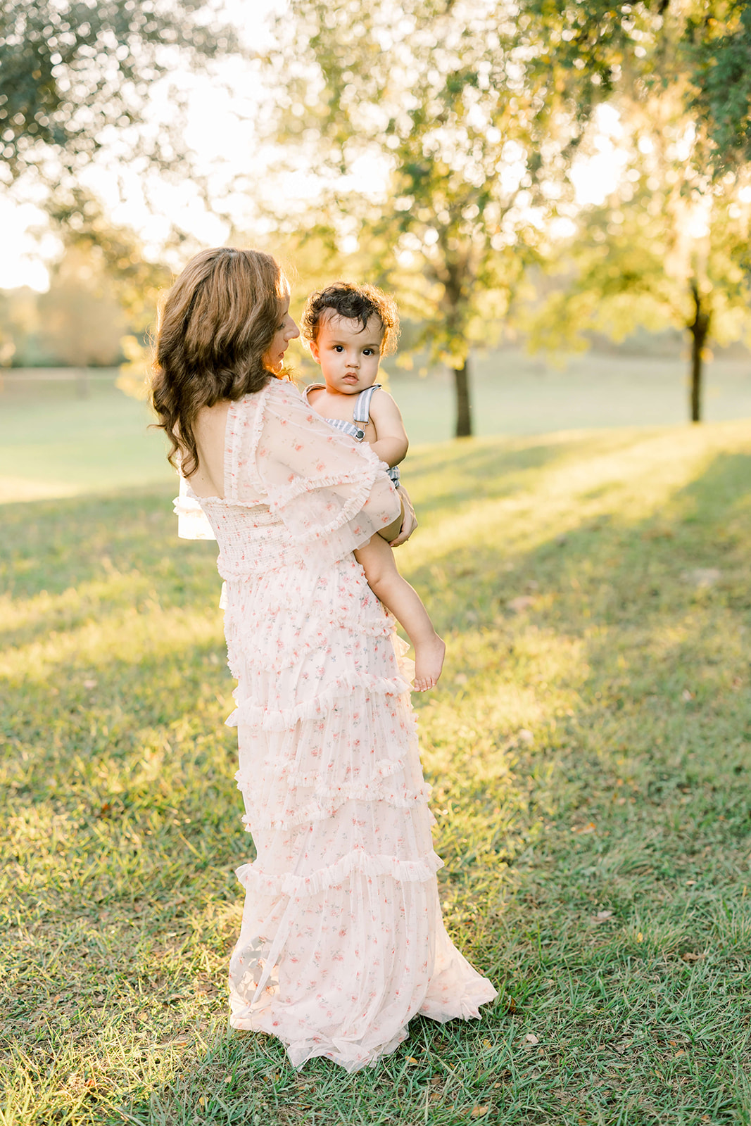 A mother in a pink dress stands in a park at sunset holding her toddler son in striped overalls before visiting Toy Stores In Houston