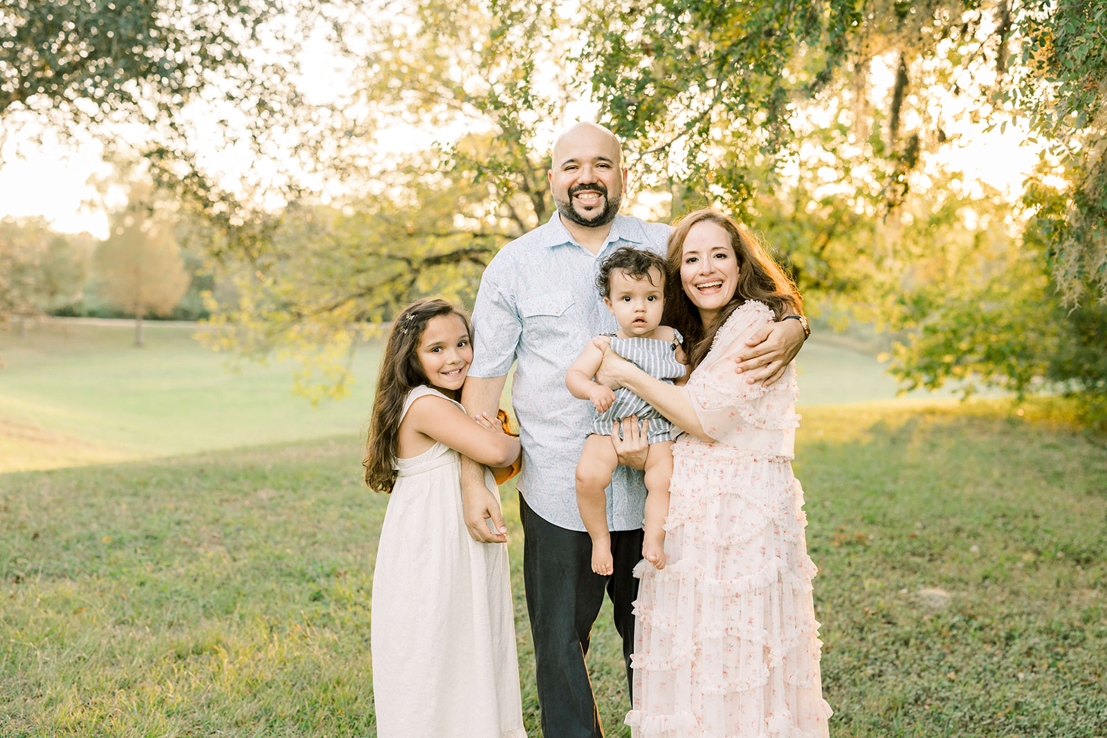 A happy mom and dad stand in a park under trees hugging their toddler son and young daughter before visiting Toy Stores In Houston